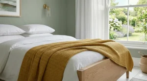 A detailed close-up photograph of a mustard yellow cotton waffle-weave throw draped over the foot of a light wooden bed frame in a natural daylight UK bedroom.