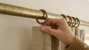 A detailed product close-up of a decorative textured brass curtain pole bracket mounted on a light wall, with matching brass rings holding linen-weave curtains.